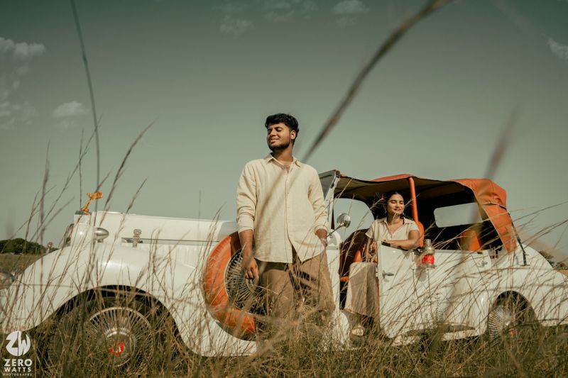 Artistic outdoor portrait with couple standing beside vintage white automobile in tall grass at sunset.