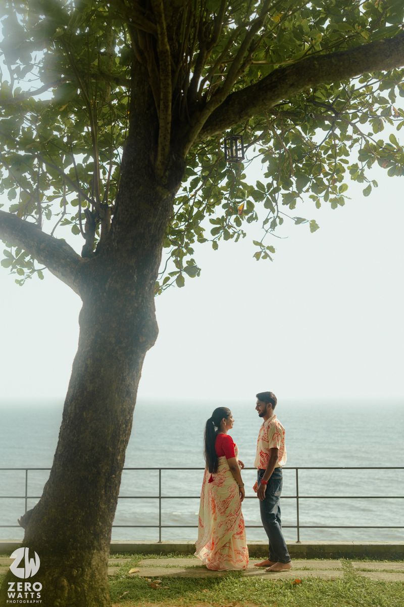 A couple shares a tender moment by the ocean under a large tree with branches creating a natural canopy.
