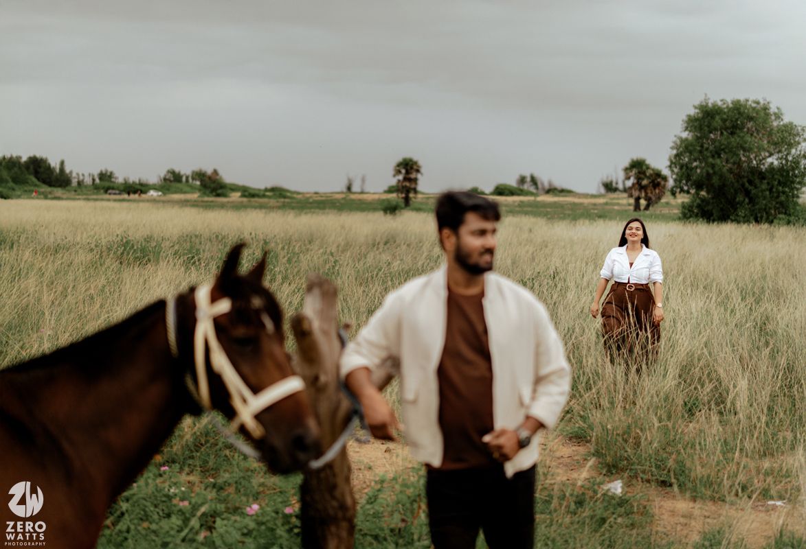 A couple stands in a grassy field with a brown horse on a cloudy day, creating a romantic countryside scene.