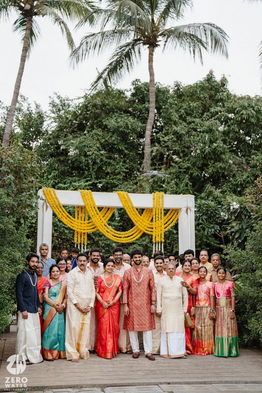 Wedding party group photo with guests in colorful traditional attire under palm trees and yellow decorations.