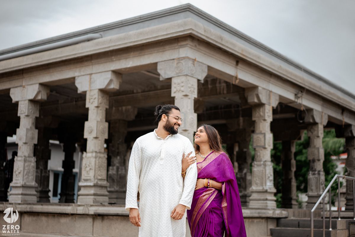 A couple in traditional Indian attire share romantic moments under ornate stone pillars of a temple courtyard.