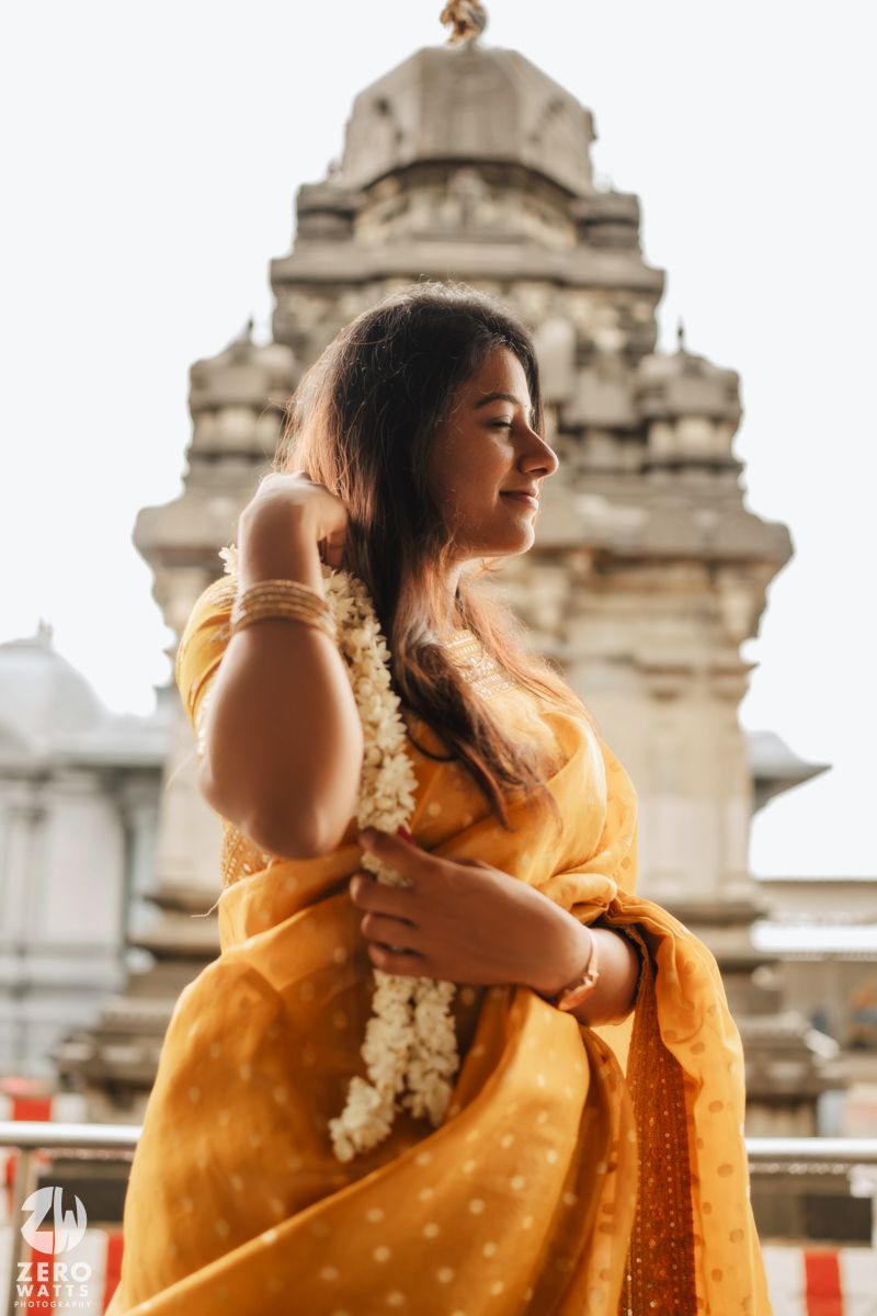 Woman in yellow traditional Indian saree poses elegantly against backdrop of ornate temple architecture.