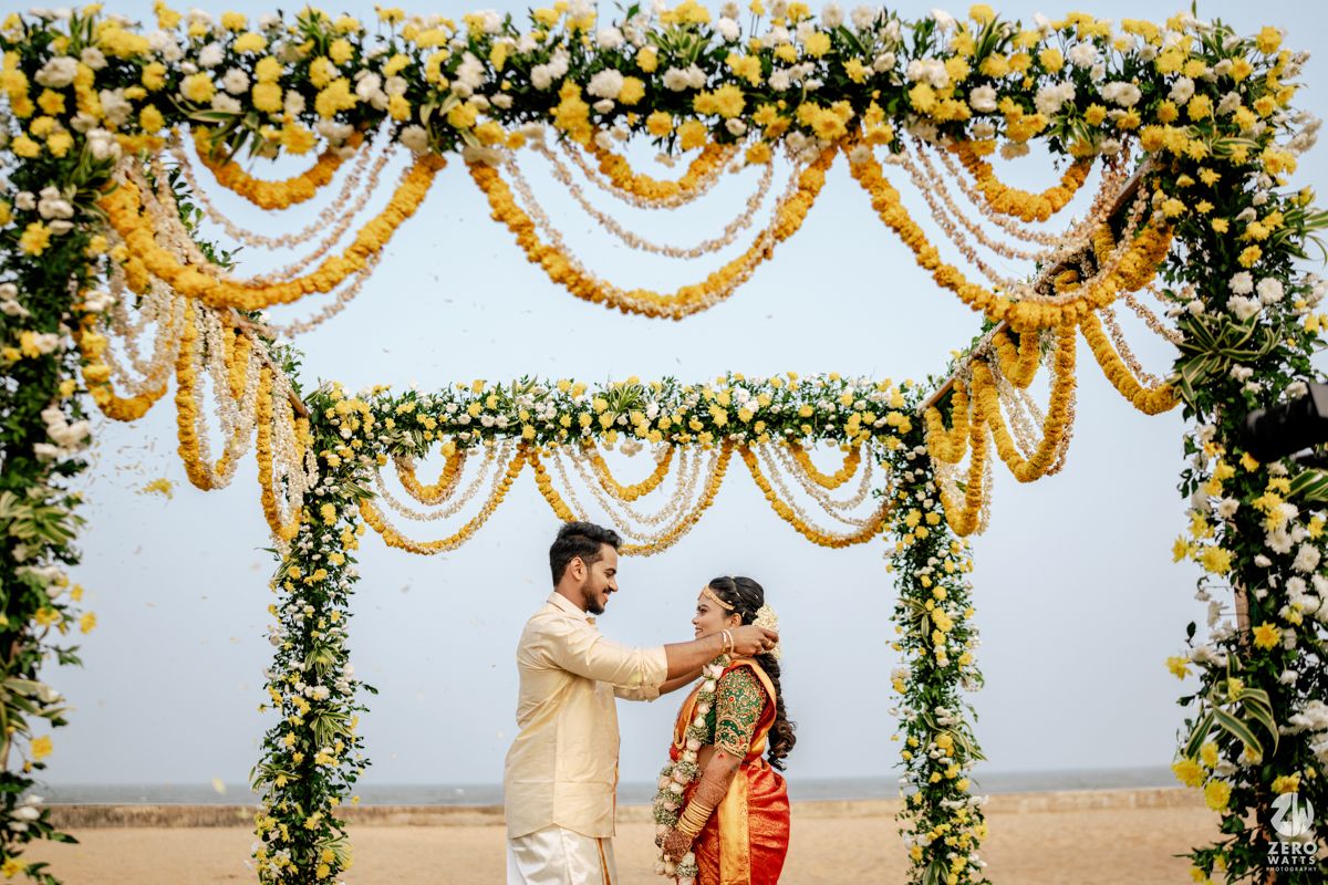 Groom places garland on bride under a vibrant yellow rose and marigold decorated mandap at Indian wedding.