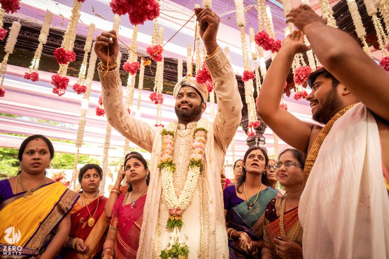 Groom in white sherwani lifted by groomsmen during vibrant South Indian wedding ritual.