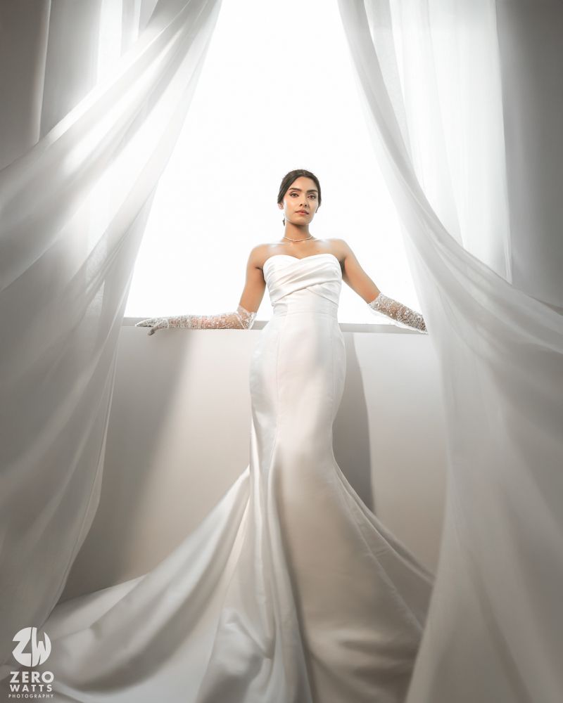 Elegant bride in a white strapless satin gown with gloves, posed between sheer curtains in soft backlight.