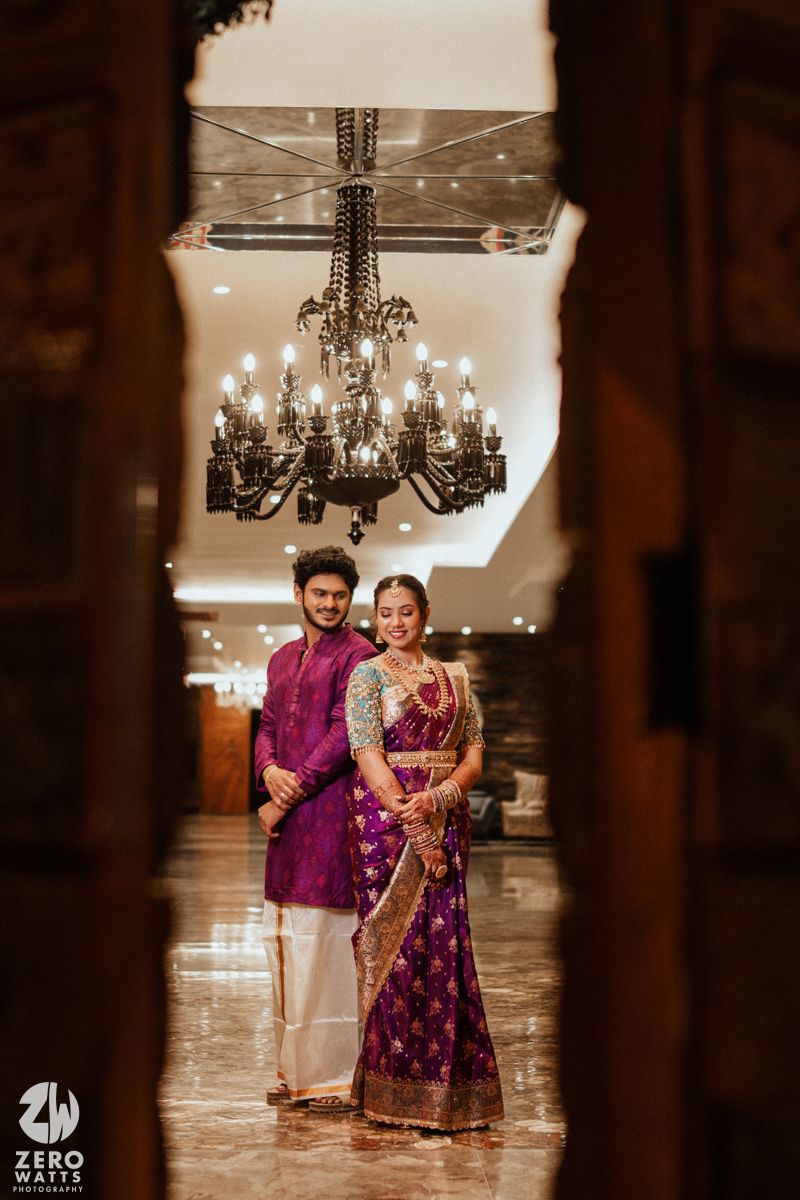 Couple in matching purple outfits posing under chandelier in elegant wedding venue corridor.