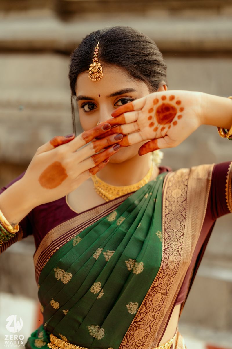 Bride in green Kanjivaram silk saree with gold jewelry displaying intricate henna mehndi designs on her hands.