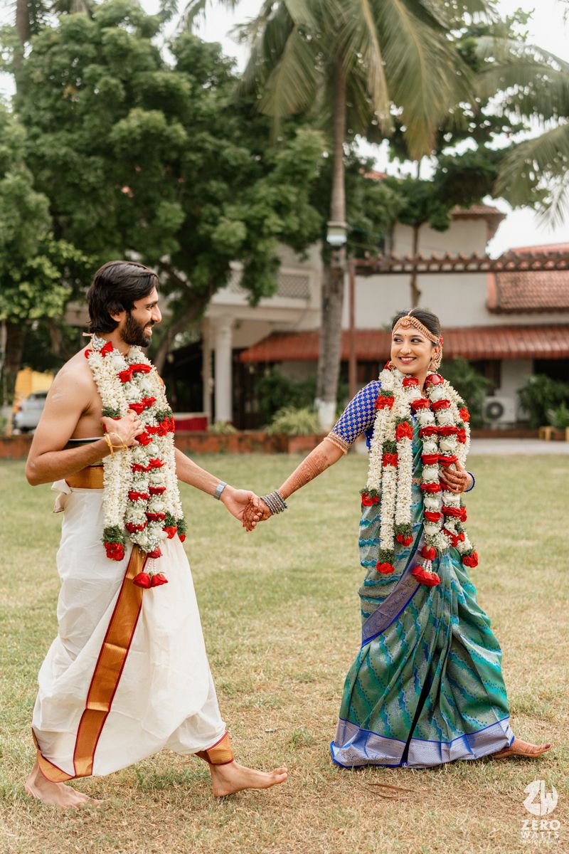 Smiling South Indian newlyweds hold hands walking outdoors wearing floral wedding garlands in traditional attire.