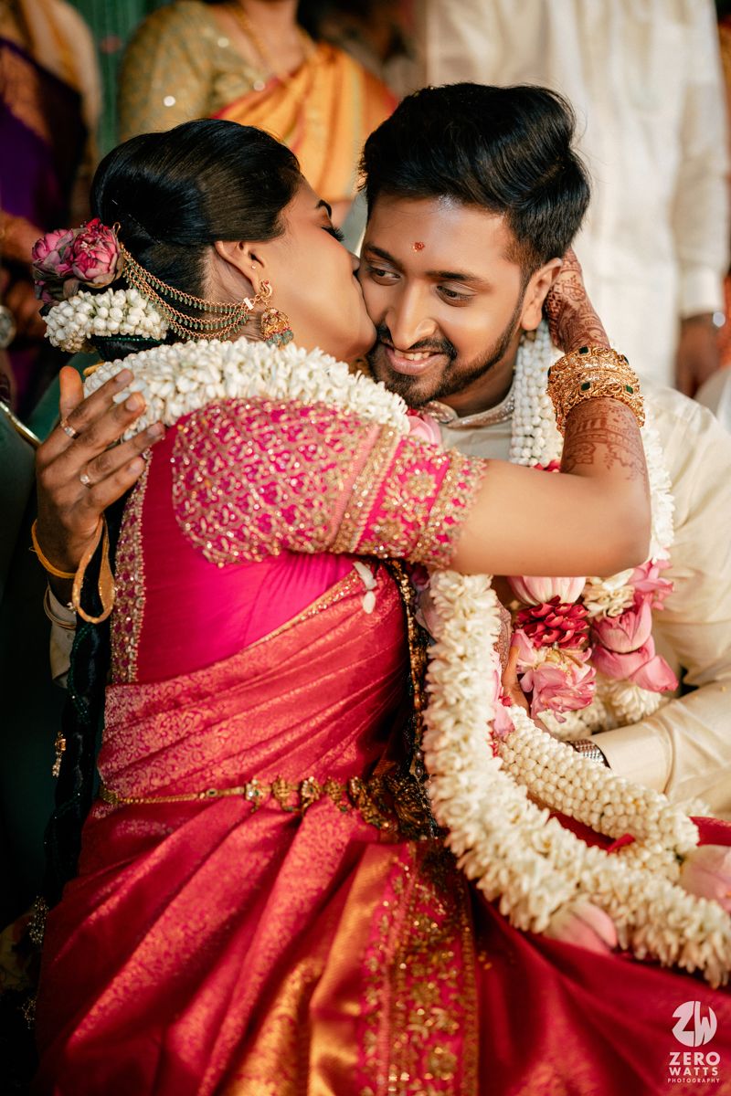 Bride in red silk saree embraces groom wearing floral garlands during South Indian wedding ceremony.