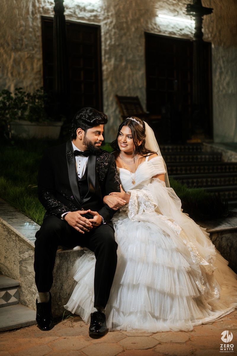 Bride and groom in elegant wedding attire sit together on steps in a romantic nighttime portrait.