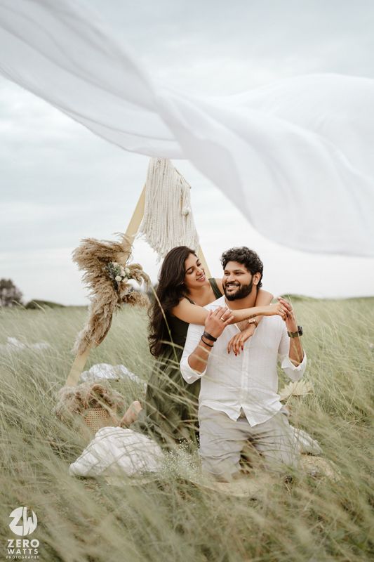 A romantic photoshoot in a windy field with flowing white fabric and tall grass creating a dreamy atmosphere.