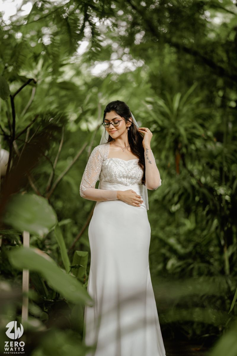 A bride in a white lace wedding dress poses thoughtfully in a lush garden setting with soft natural lighting.