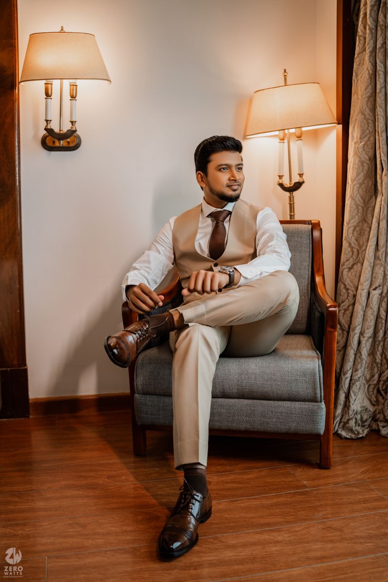 Stylish groom in beige suit poses on grey armchair in elegant room with warm lighting and hardwood floors.