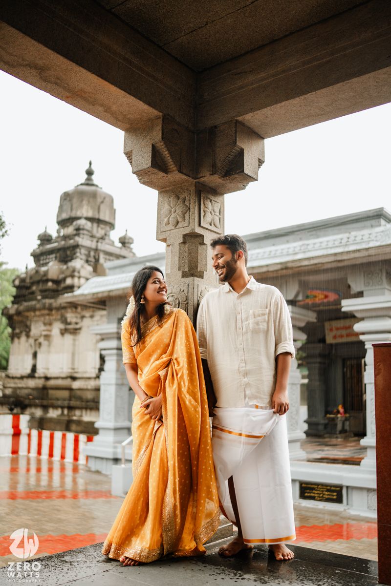 Couple in white and orange outfits posing together at a traditional temple with stone architecture.