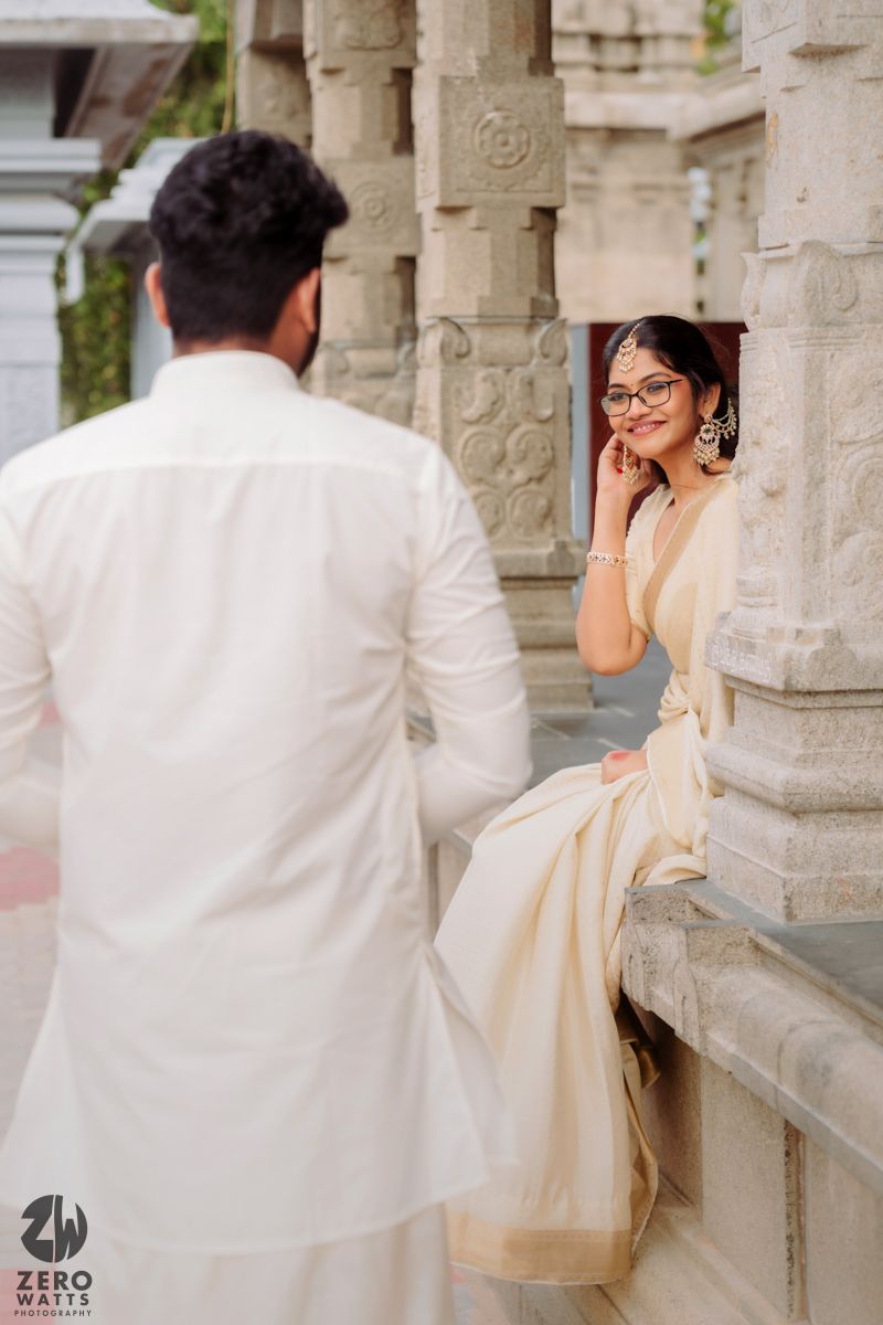 A romantic couple in white and cream outfits pose together in an elegant stone colonnade setting.