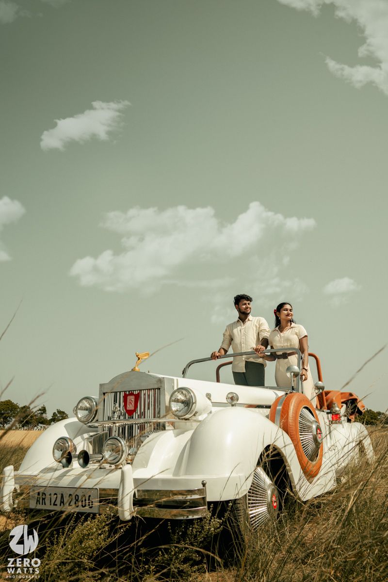 A couple poses atop a pristine white vintage car in a field during a retro-styled photoshoot.