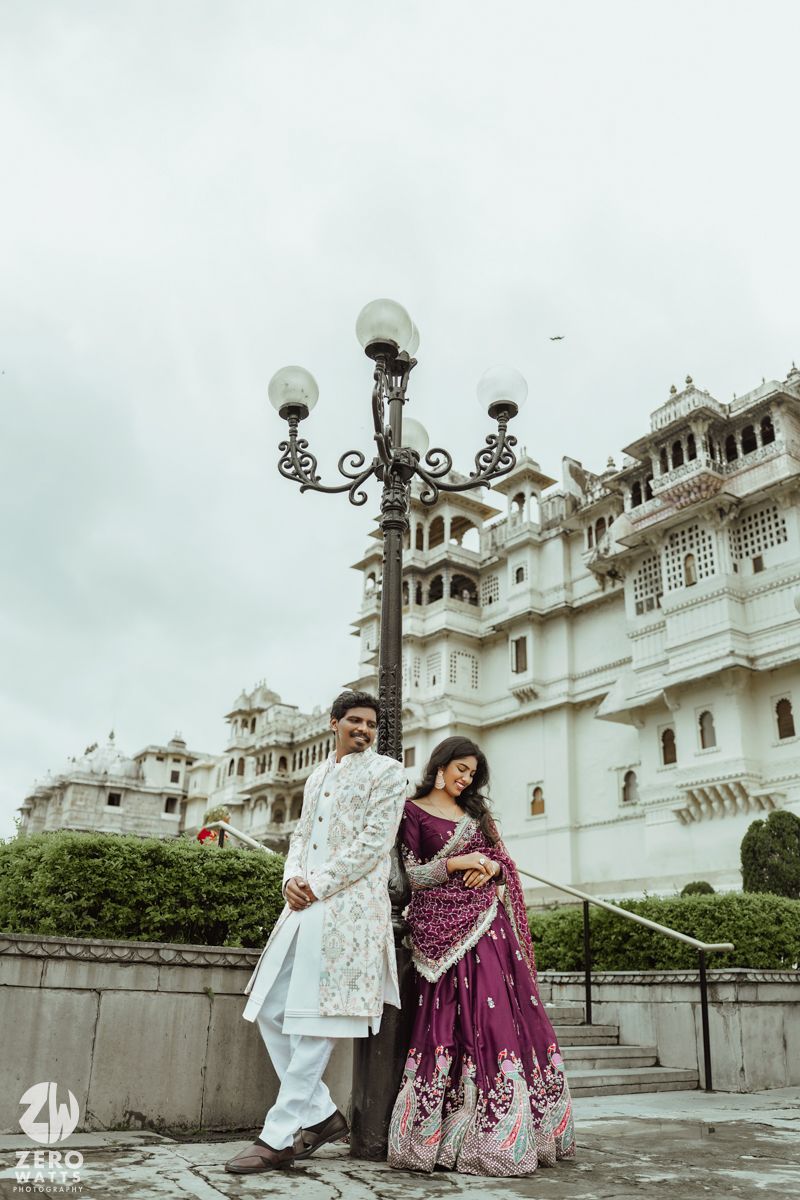 A couple in elegant Indian attire poses near ornate white palace architecture with vintage-style lampposts.