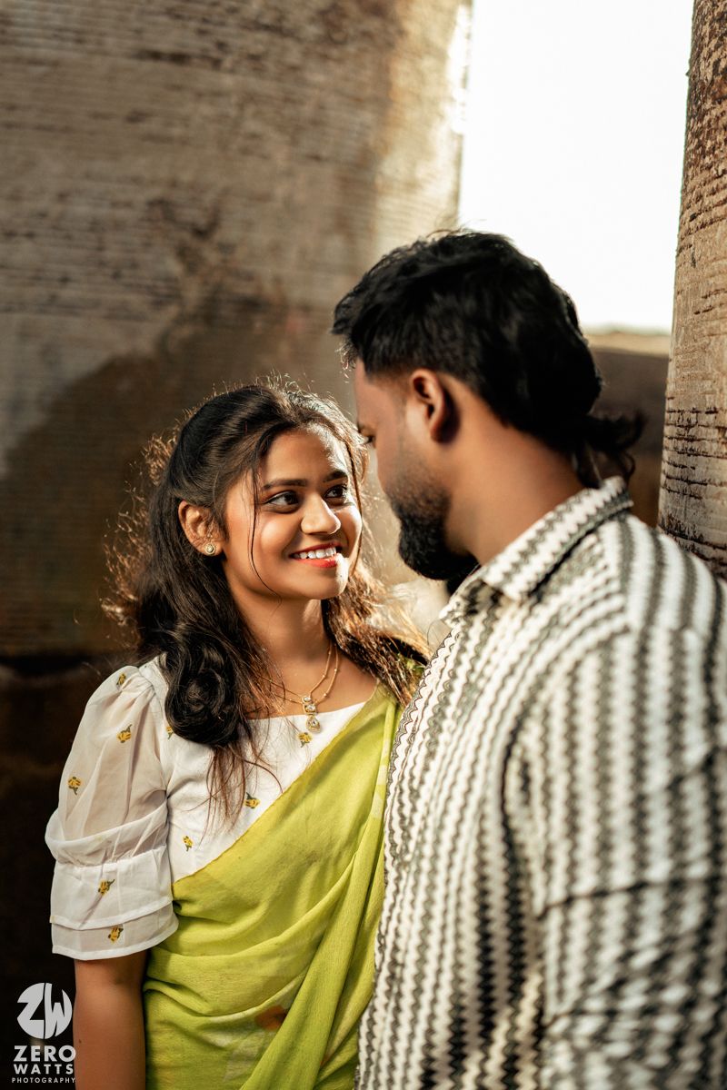 A couple shares an intimate moment against a brick wall backdrop, with sunlight creating a warm glow.