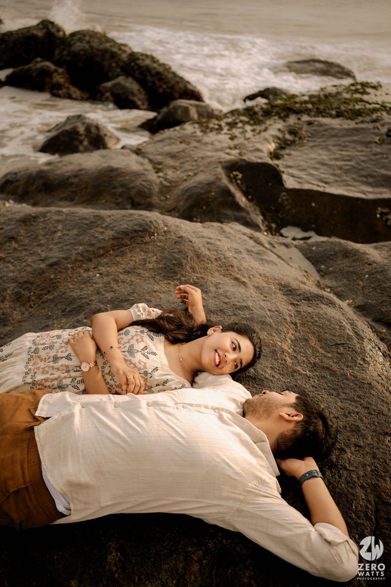 Couple lying on coastal rocks at sunset in romantic beach photoshoot wearing white clothing.