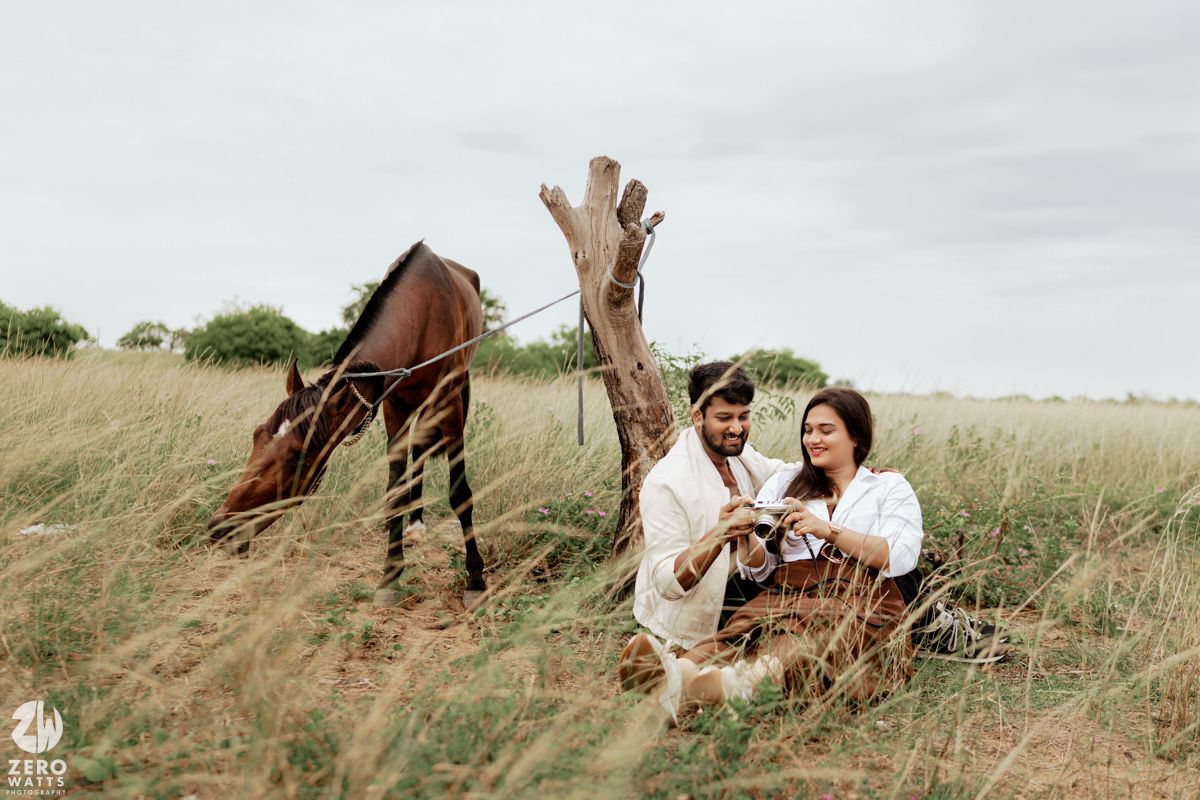 A couple sits together near a weathered tree trunk while their horse grazes in a grassy field.
