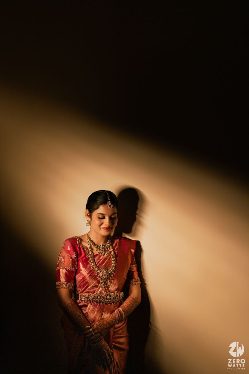 South Indian bride in pink silk saree and gold jewelry posed under dramatic spotlight against dark wall.