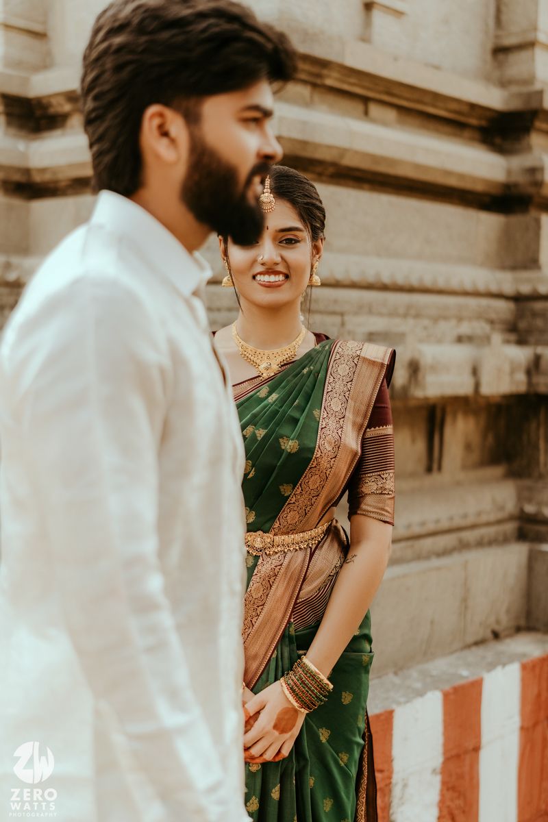 South Indian bride in green silk saree smiling at groom in white kurta during traditional wedding photoshoot.