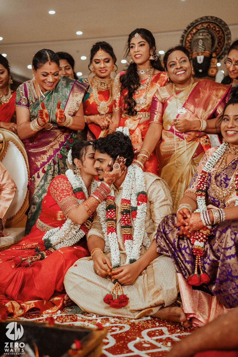 South Indian groom and bride sit during wedding ritual surrounded by smiling family members in silk sarees and jewelry.