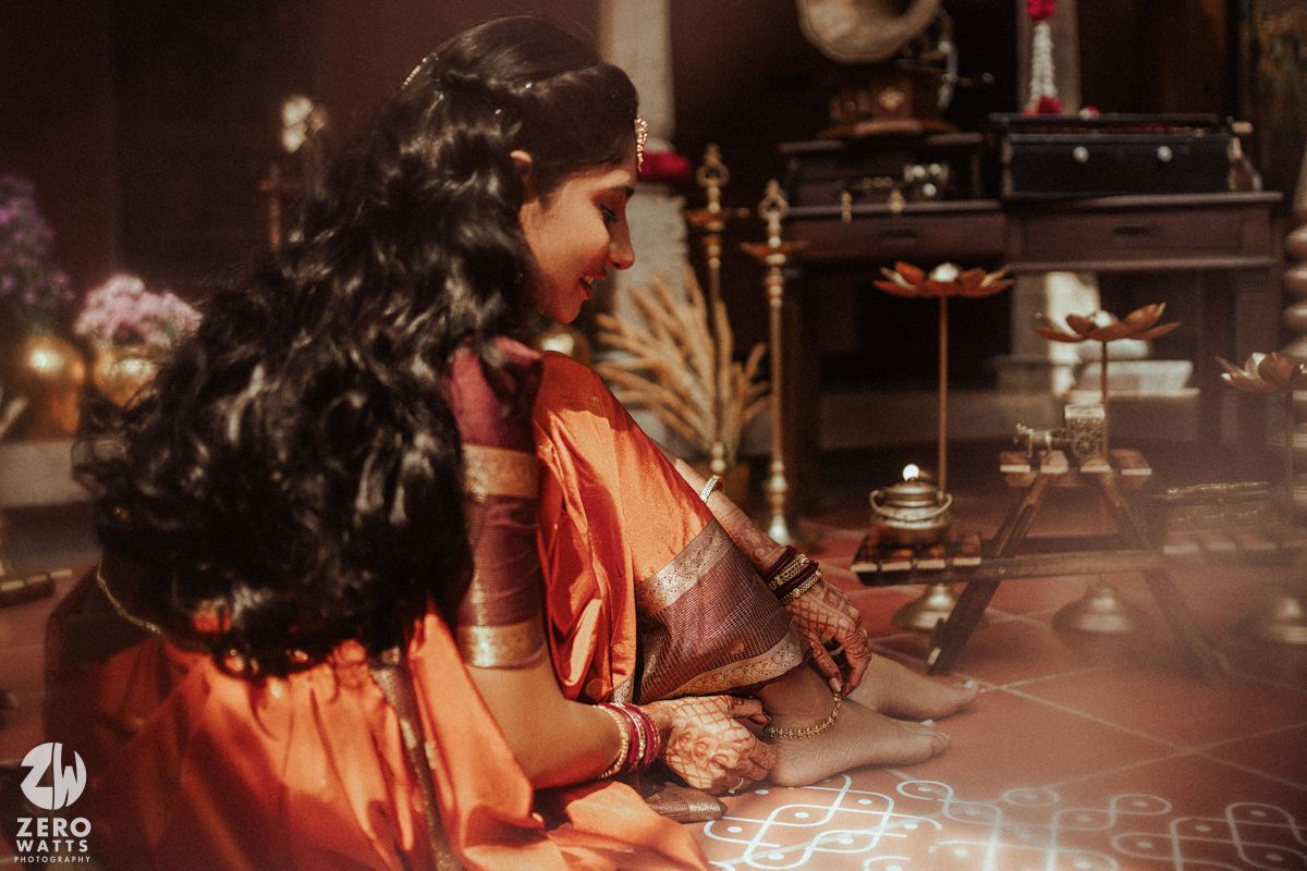 Two women in traditional orange silk sarees participate in a South Indian Hindu ritual ceremony indoors.