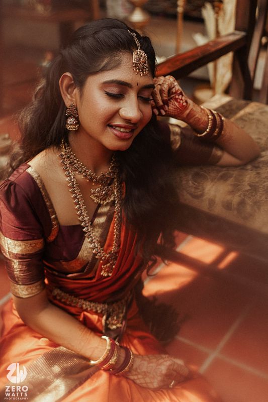 Beautiful South Indian bride adorned with gold jewelry, maang tikka, and mehndi in orange Kanjivaram saree.