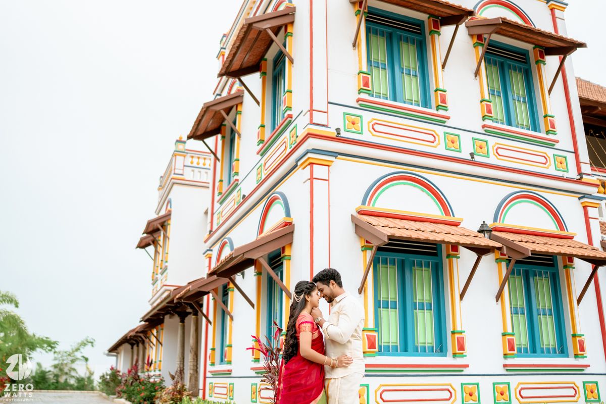 Couple in traditional Indian attire embracing before a colorful multi-story building with vibrant architectural details.