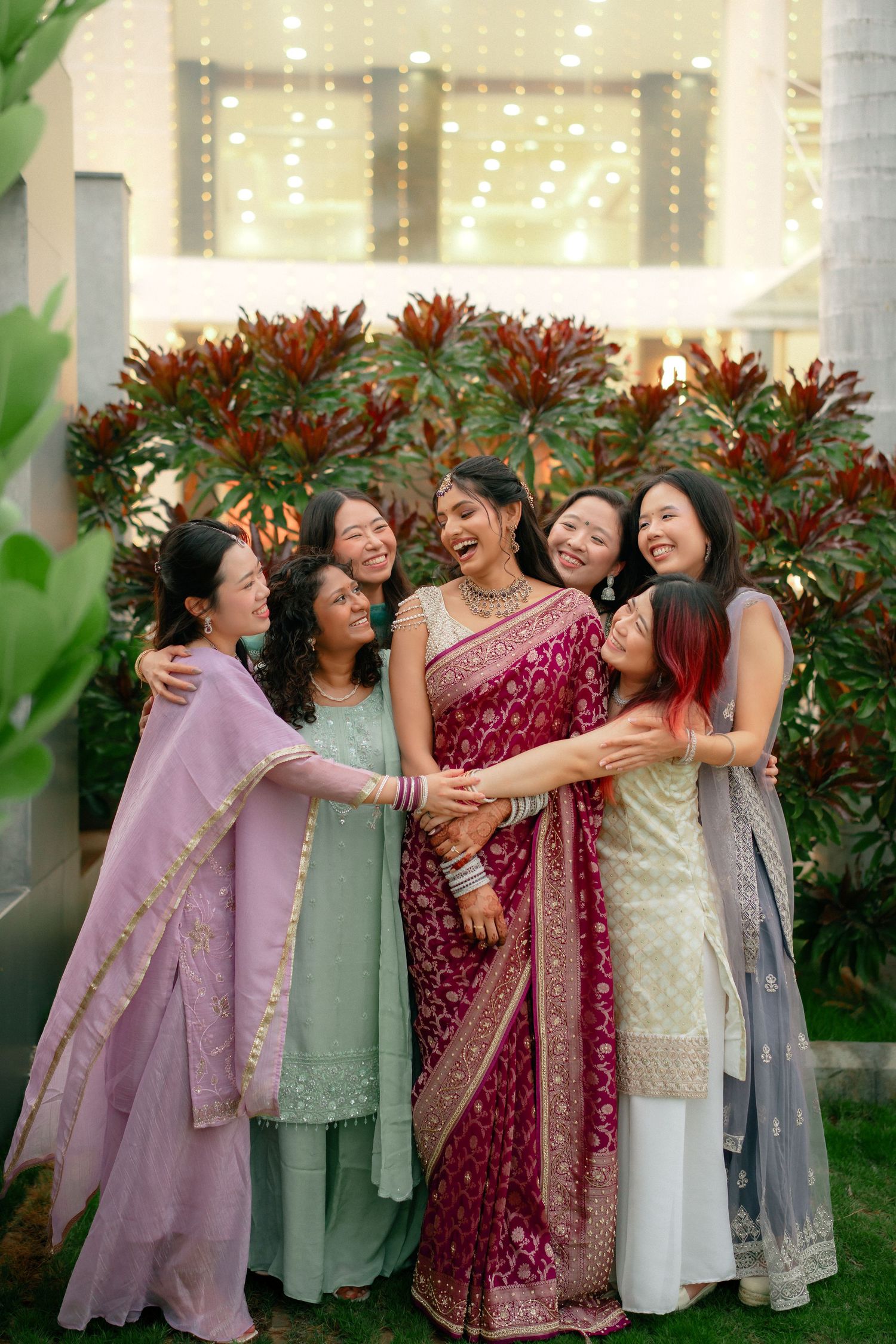 Group of smiling women in colorful traditional Indian saris and salwar suits embracing outdoors in front of red-leaved plants.