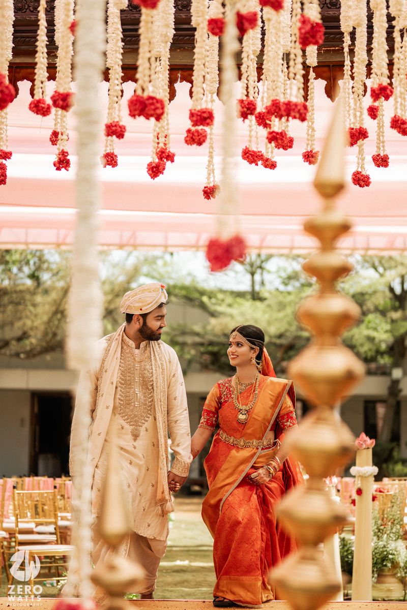 Indian couple in traditional attire holding hands under floral decorations at a South Indian wedding ceremony.