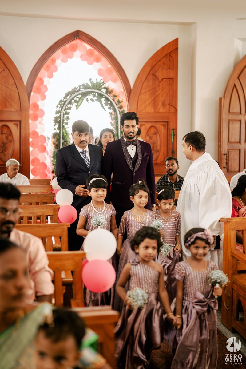 Groom walks down church aisle with flower girls holding pink balloons at a Christian wedding ceremony.