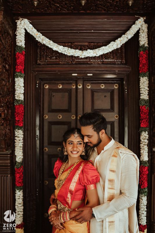 South Indian wedding couple embracing before ornate carved wooden door decorated with white and red garlands.