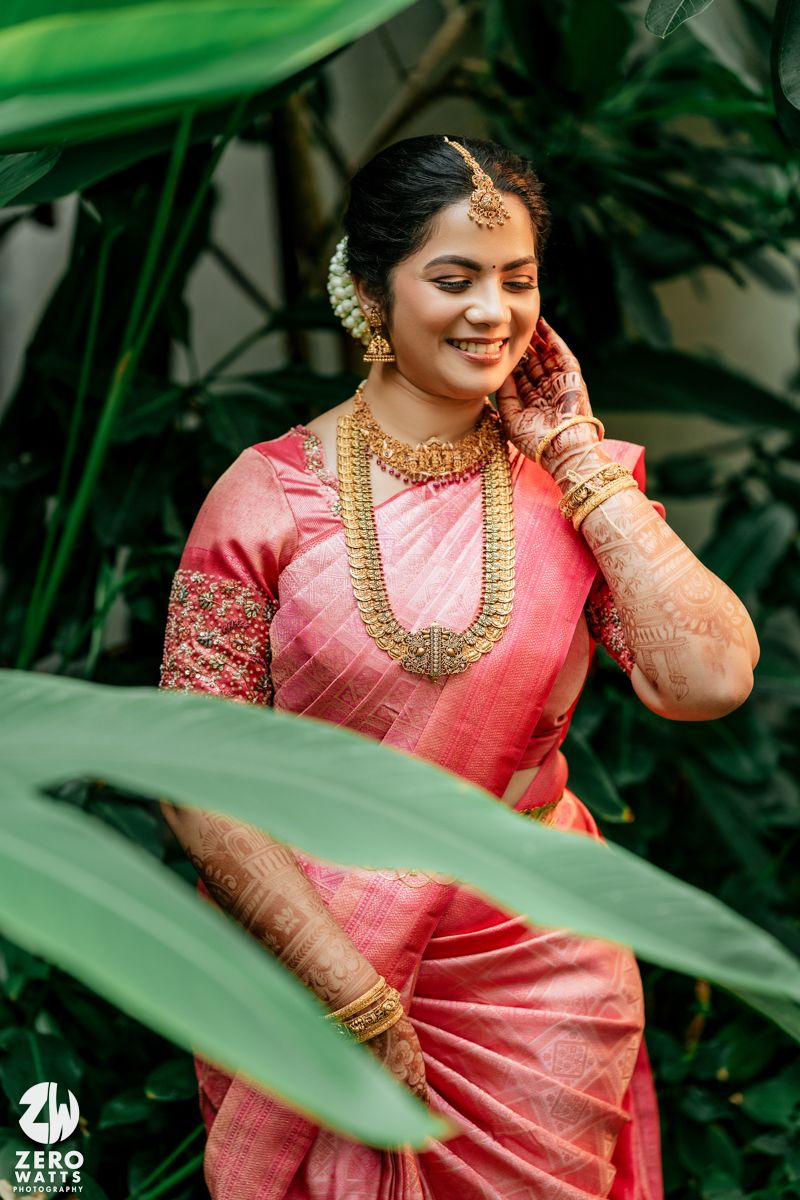 South Indian bride in pink silk saree with gold jewelry and mehndi smiling outdoors.