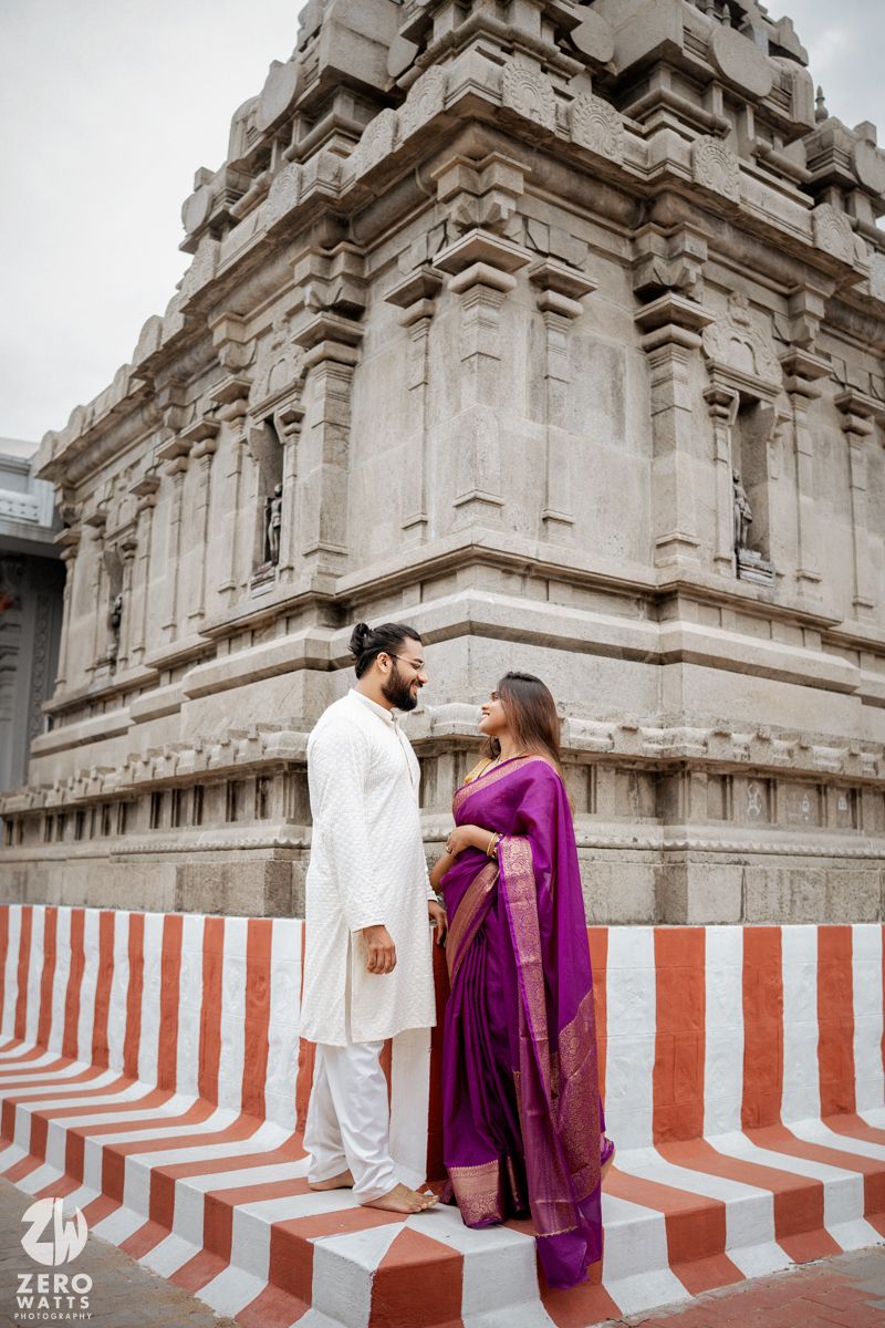 Couple in traditional attire standing against red and white striped temple walls.