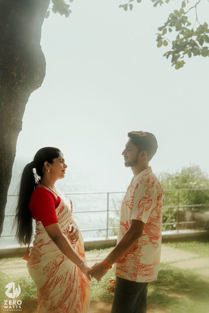 A couple in matching red and white outfits share a tender moment under a tree by the sea.