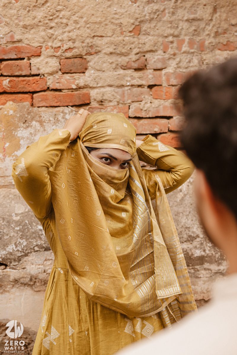 Person in flowing golden dress poses dramatically against rustic brick wall background.