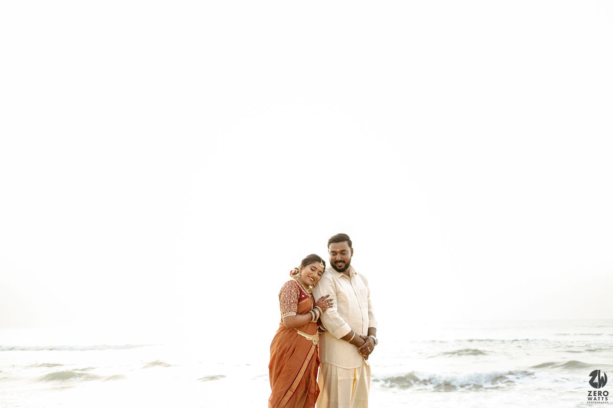 A couple in traditional Indian wedding attire poses together on a sunny beach near the ocean waves.