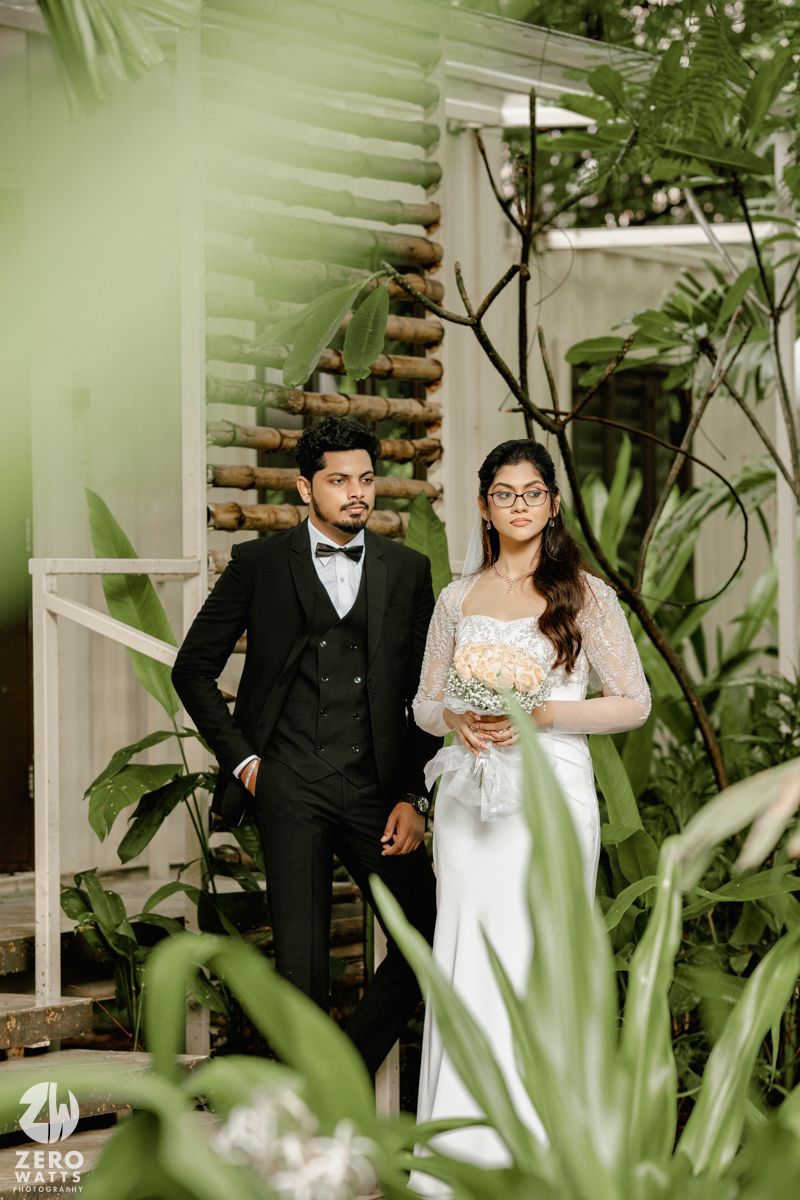 A couple poses together in an elegant garden setting surrounded by lush tropical plants and white architectural details.