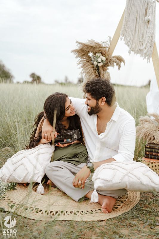 Couple enjoying romantic picnic on round woven rug with pampas grass decor and white fabric backdrop in field.