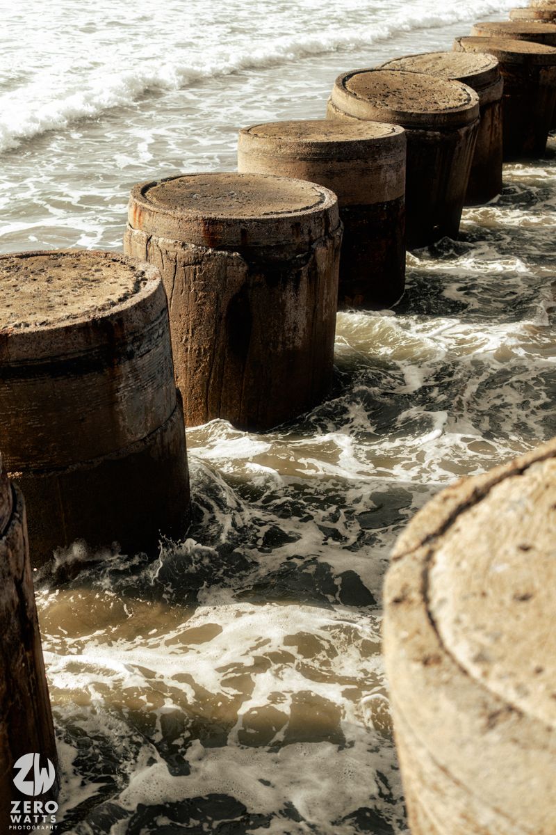 Large wooden pillars stand in rushing ocean waves, creating a dramatic coastal scene.