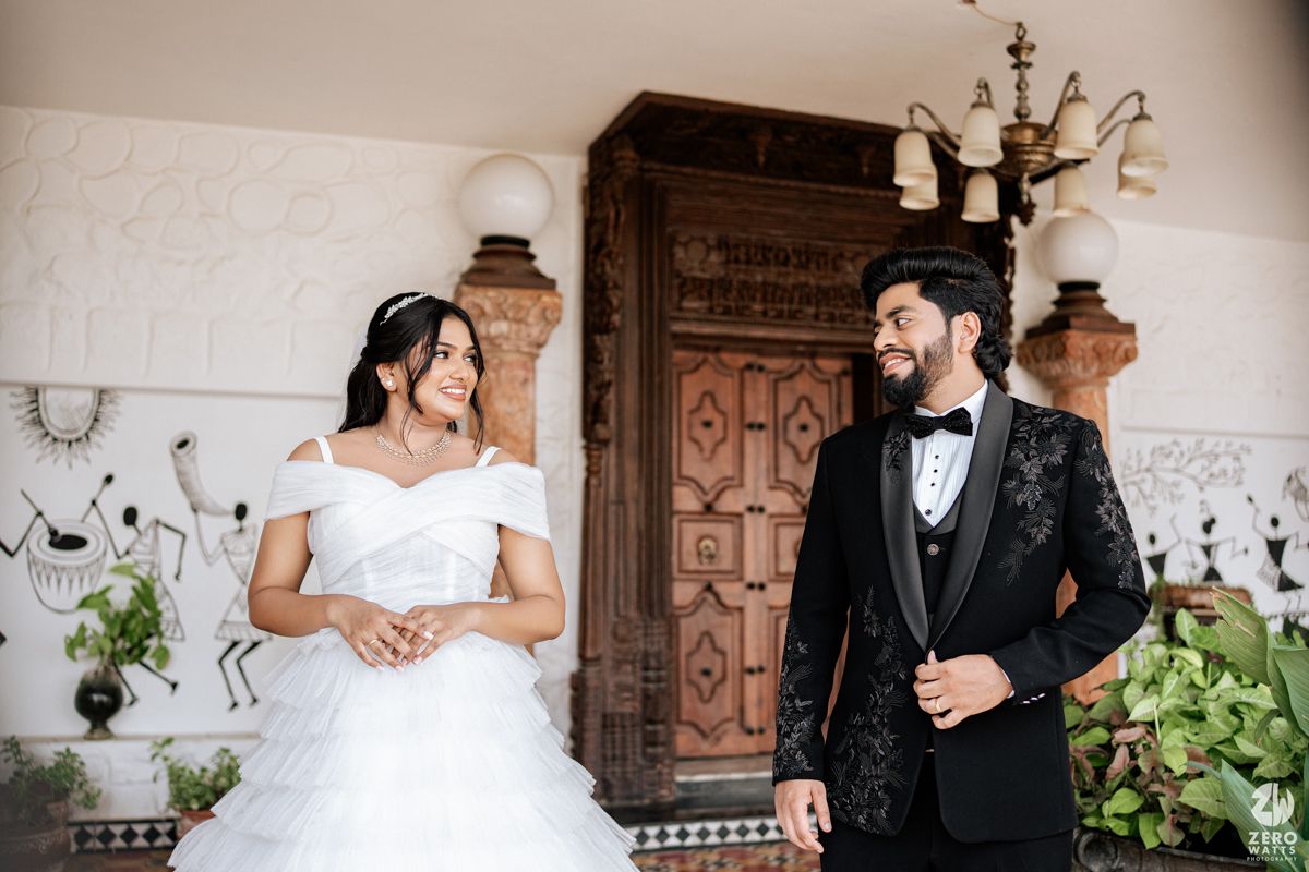 A bride in a white off-shoulder gown and groom in black tuxedo share a joyful moment outside an ornate wooden door.