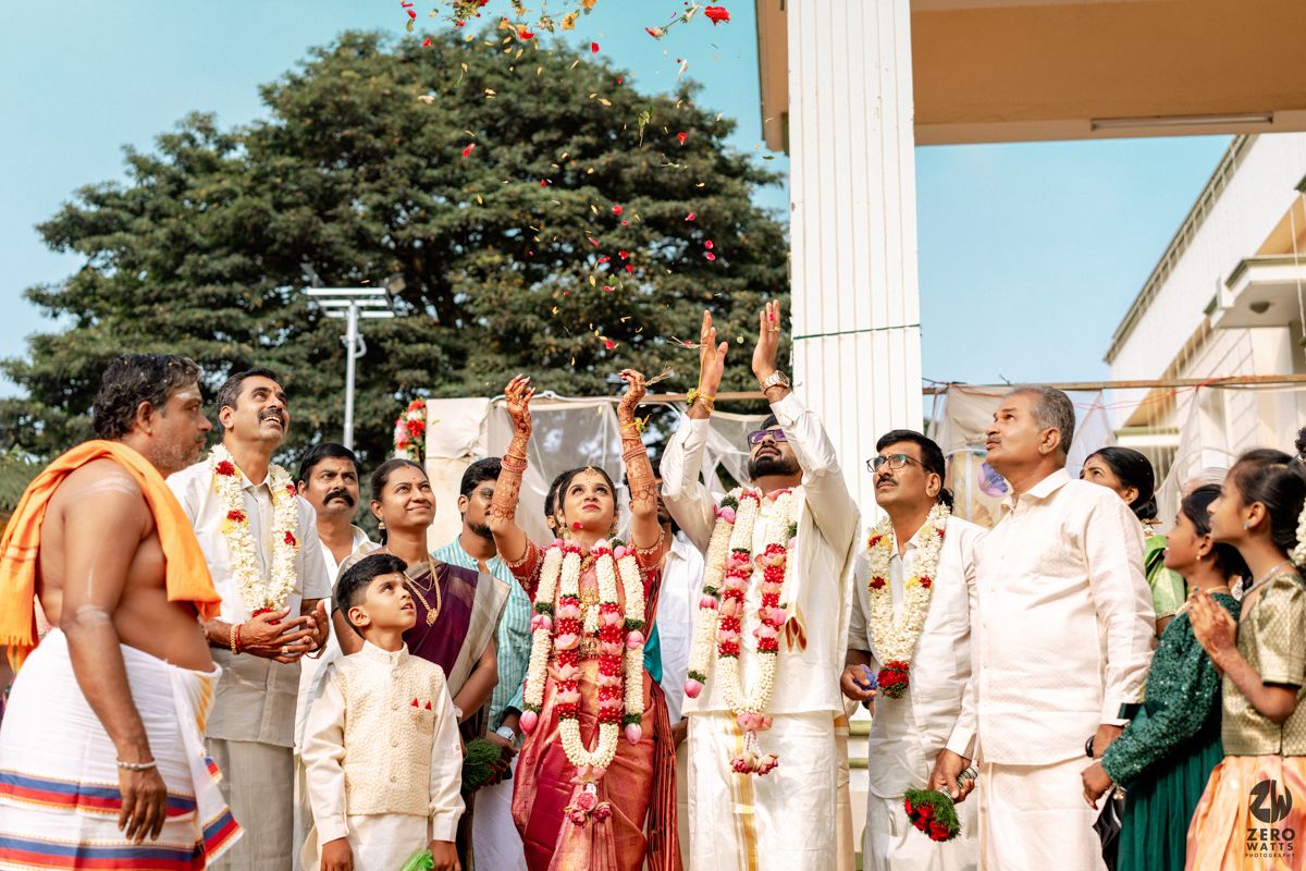 South Indian groom tossing flower petals skyward during traditional outdoor wedding ritual with guests.