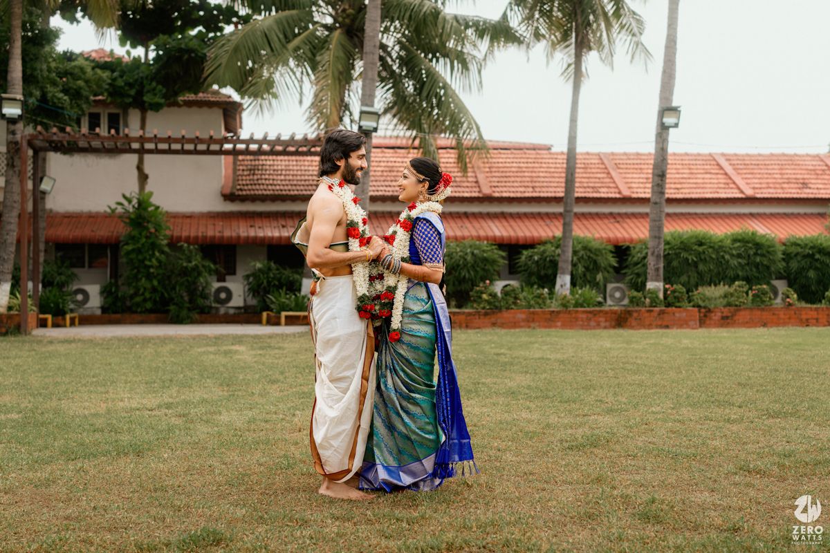 South Indian bride and groom in traditional attire stand together on green lawn with palm trees backdrop.
