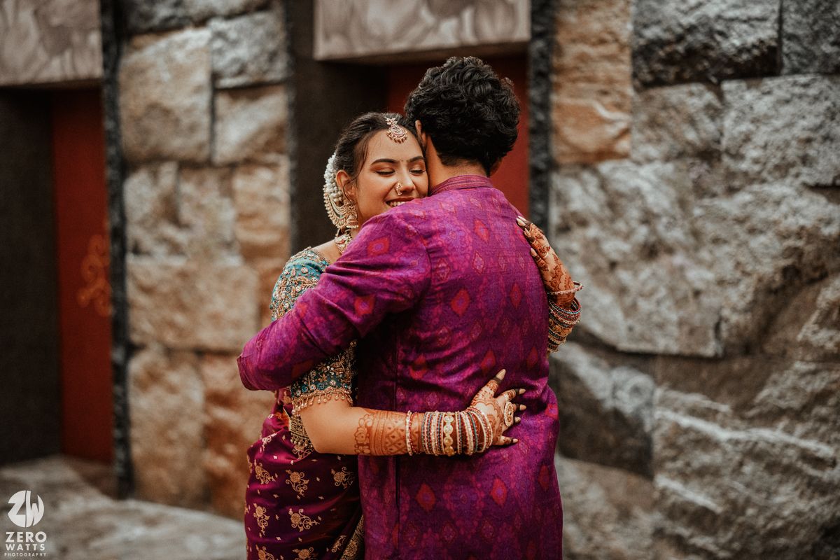Bride and groom in vibrant Indian wedding attire share a warm embrace outdoors against a stone wall backdrop.