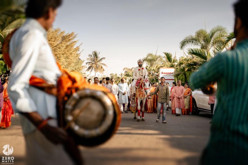 Indian groom on horse during baraat procession with dhol players and wedding guests on sunny outdoor road.