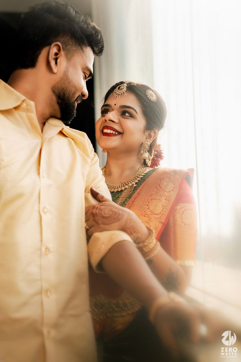 South Indian bride in red saree smiling up at groom in cream kurta in warm natural window light.
