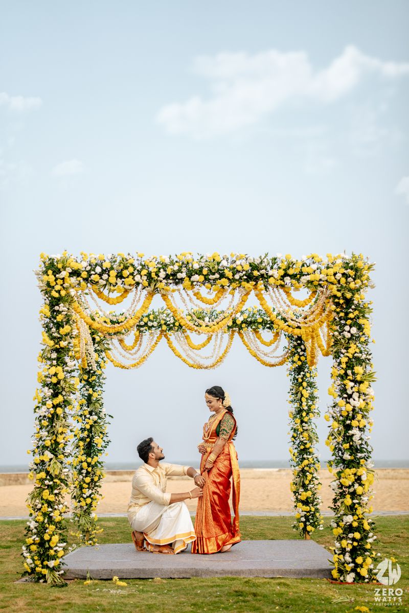Indian wedding couple under floral mandap decorated with yellow marigolds and white flowers at beach venue.