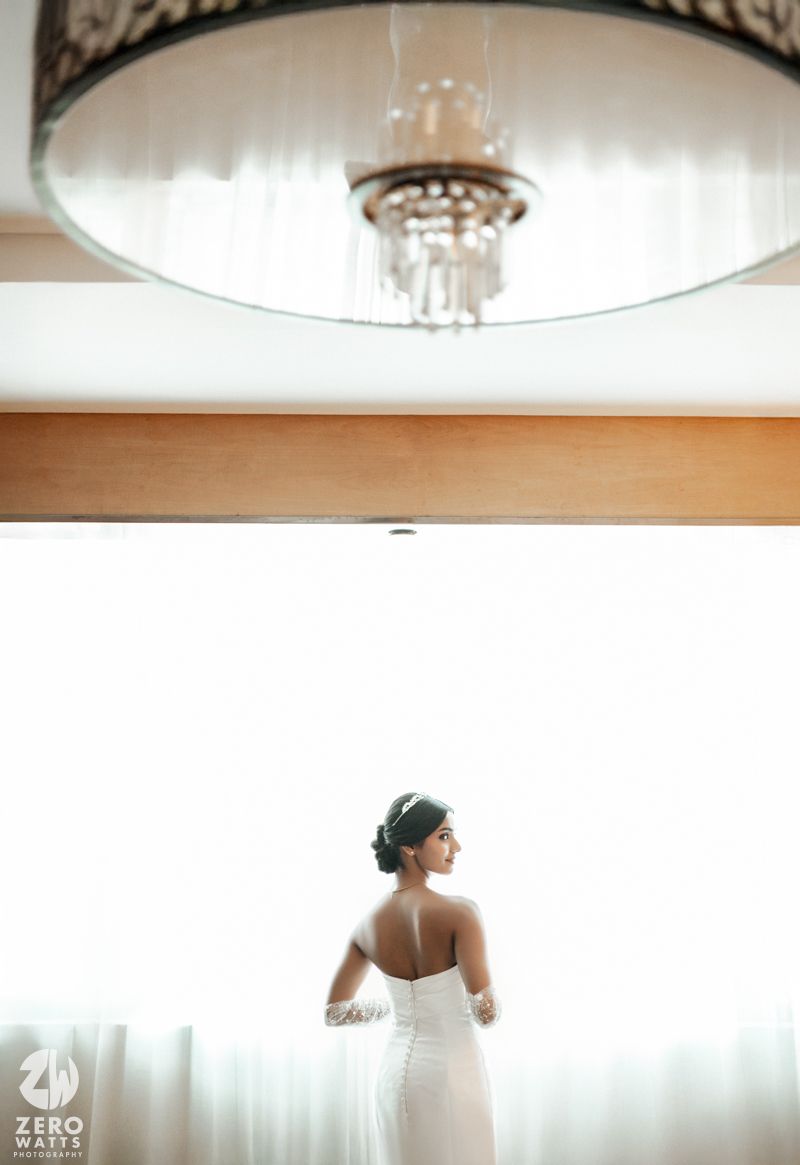 Bride in strapless white gown stands by window with chandelier above in elegant bridal suite.
